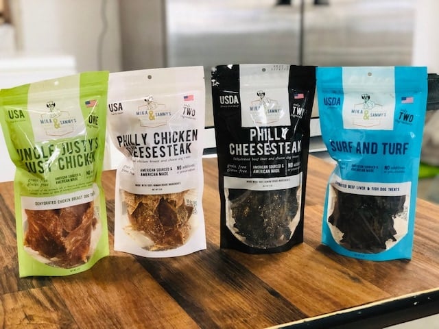 Front view of four Mika and Sammy’s pet treat pouches: Uncle Fusty’s chicken (green), Philly Chicken Cheesesteak (white and black), and Surf and Turf (blue) on a wooden counter