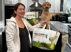 Woman holding a Mika & Sammy’s tote bag of chews with a small dog on a kitchen counter in the background
